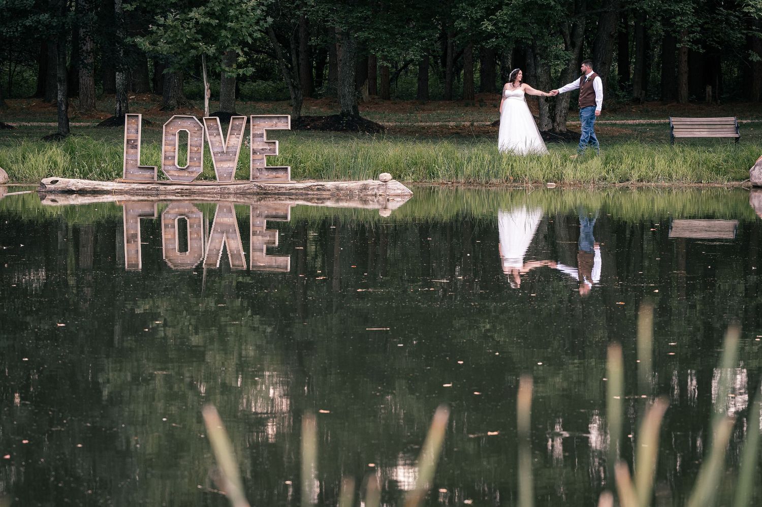 Bride and groom hold hands near large LOVE sign perfectly reflected in tranquil pond at sunset.