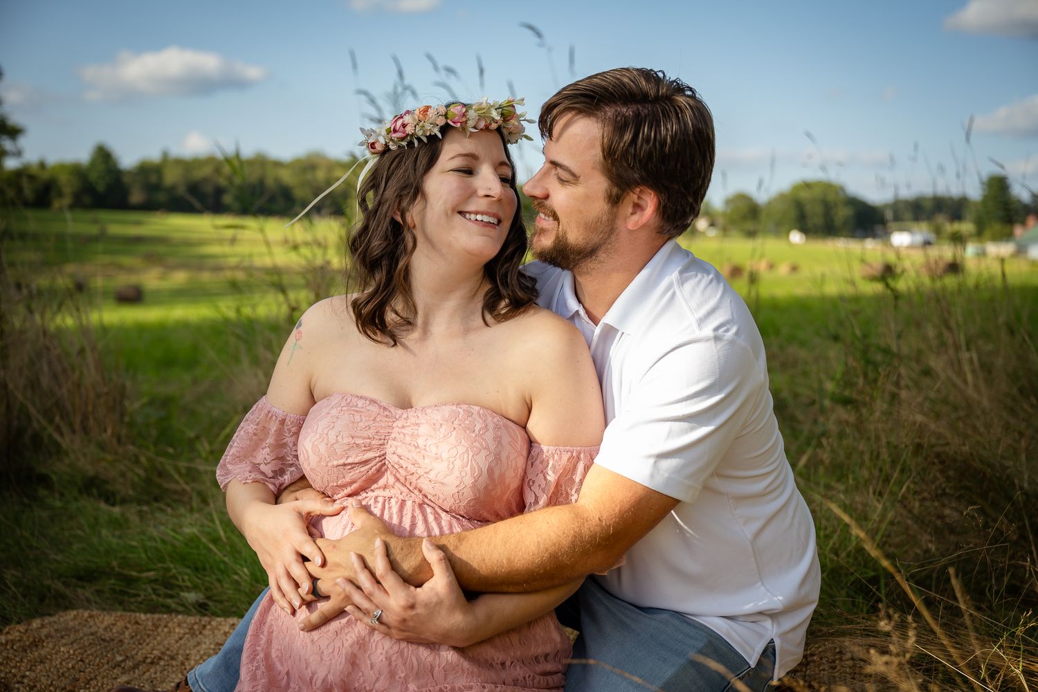 Couple sharing a tender moment outdoors, pregnant woman in pink dress with floral crown, man in white shirt embracing her.