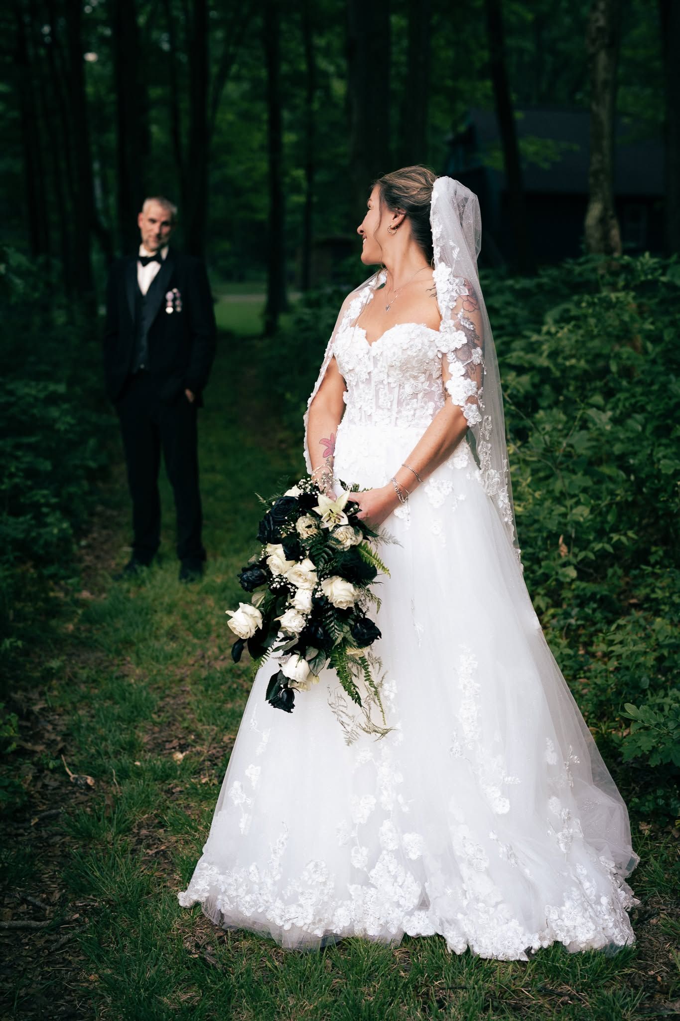Elegant bride in lace wedding gown holding bouquet stands in forest with groom in background.