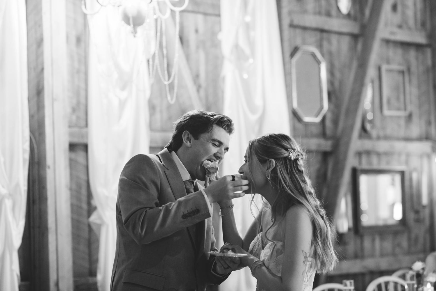 Black and white photo of bride and groom sharing wedding cake during rustic barn reception celebration.