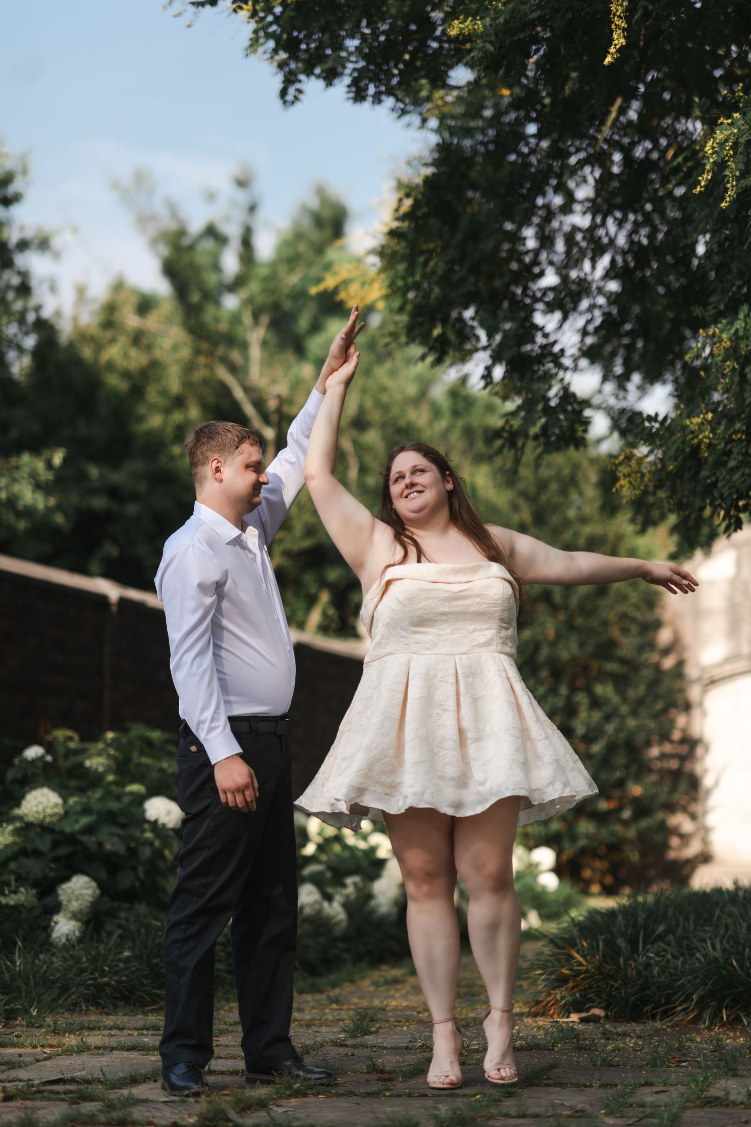 Couple dancing outdoors in garden, woman in white dress spinning with raised arm, man in white shirt guiding her.