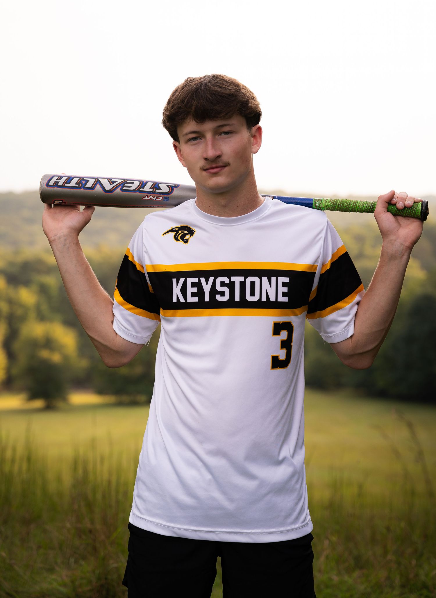Young baseball player in Keystone #3 jersey poses with bat behind shoulders in outdoor field setting.