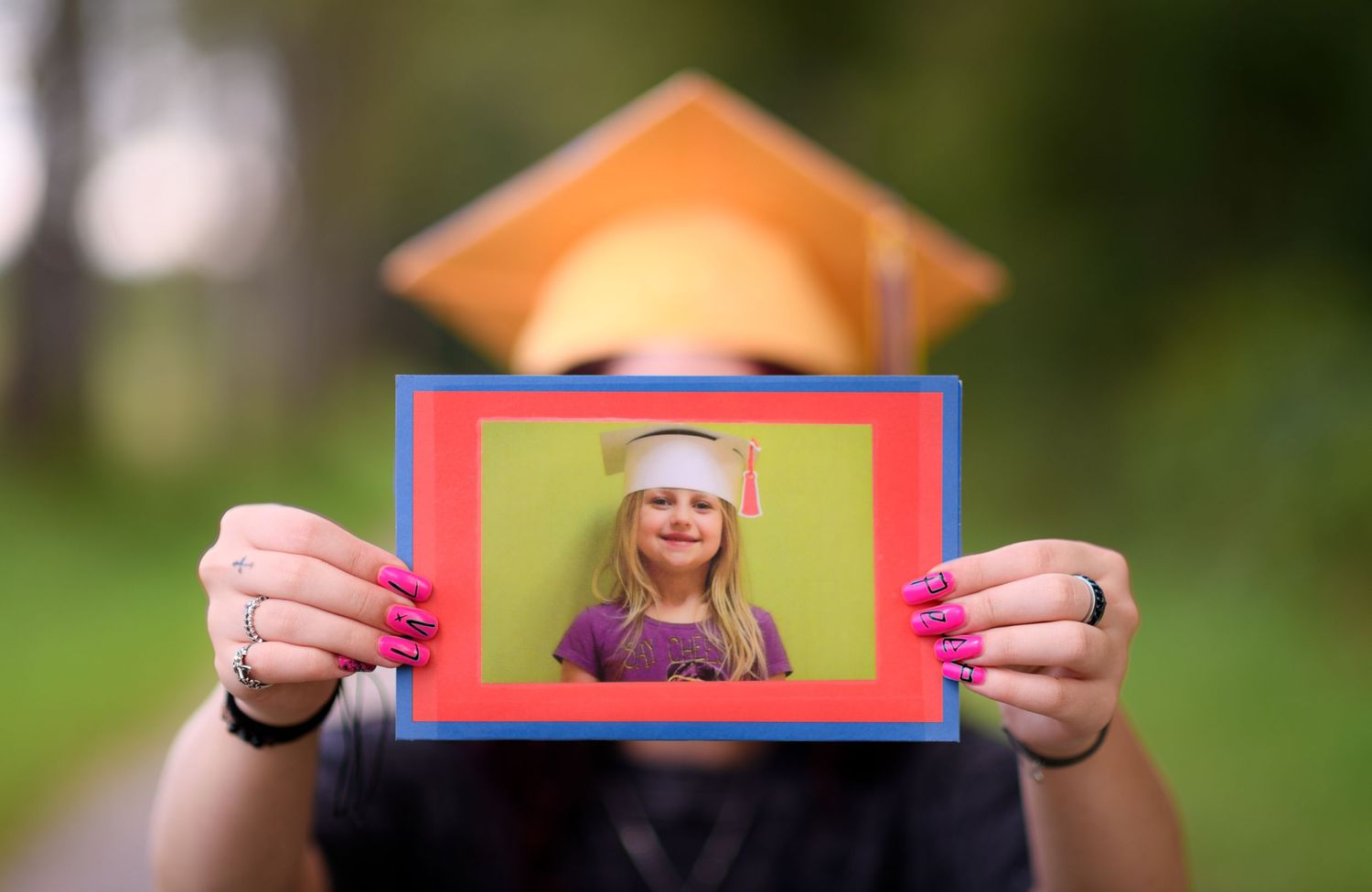 Graduate holds photo frame revealing younger self in cap, creating creative dual graduation portrait outdoors.