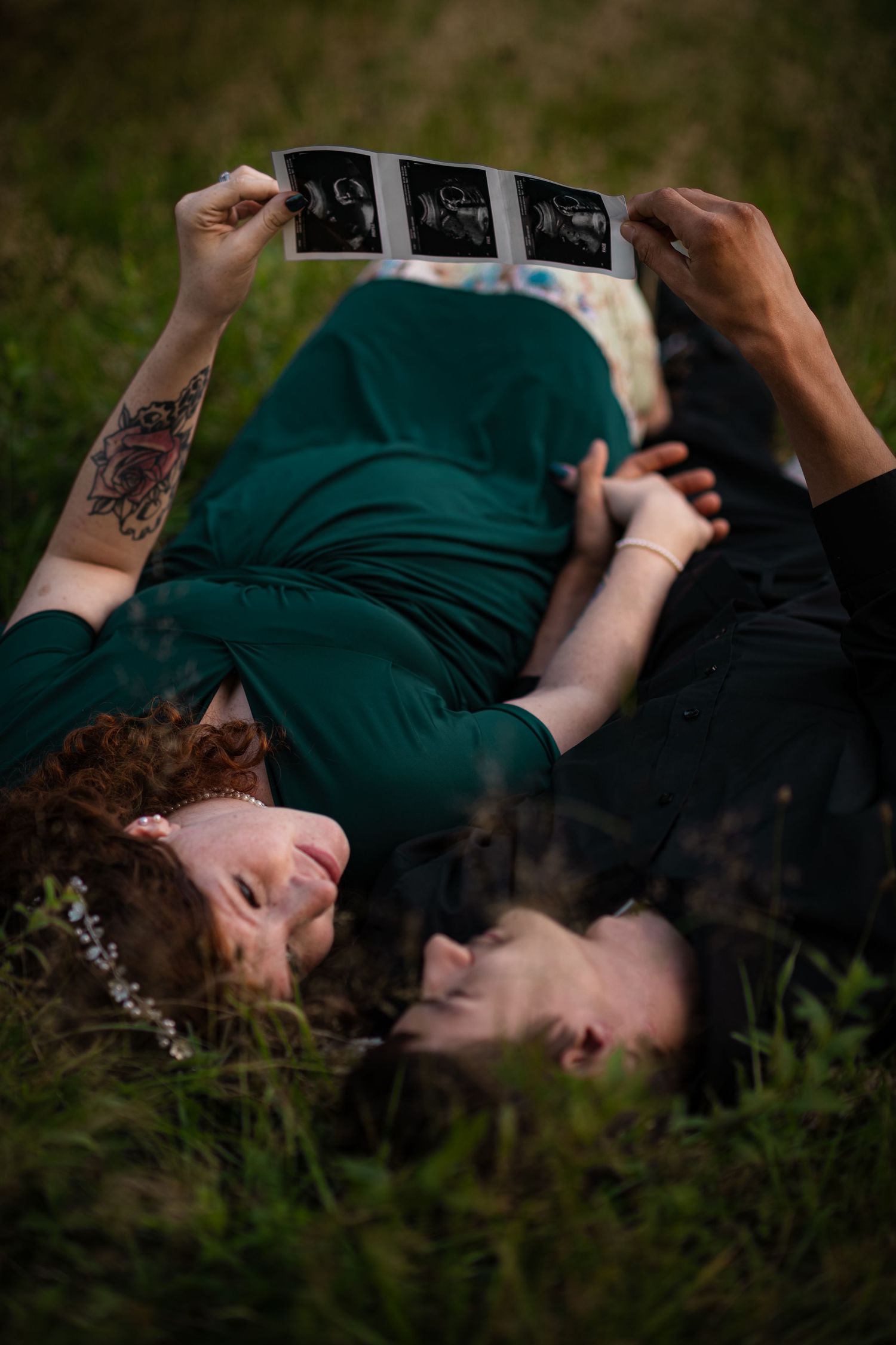 Expecting couple lies in grass holding ultrasound photos above them in a tender moment.