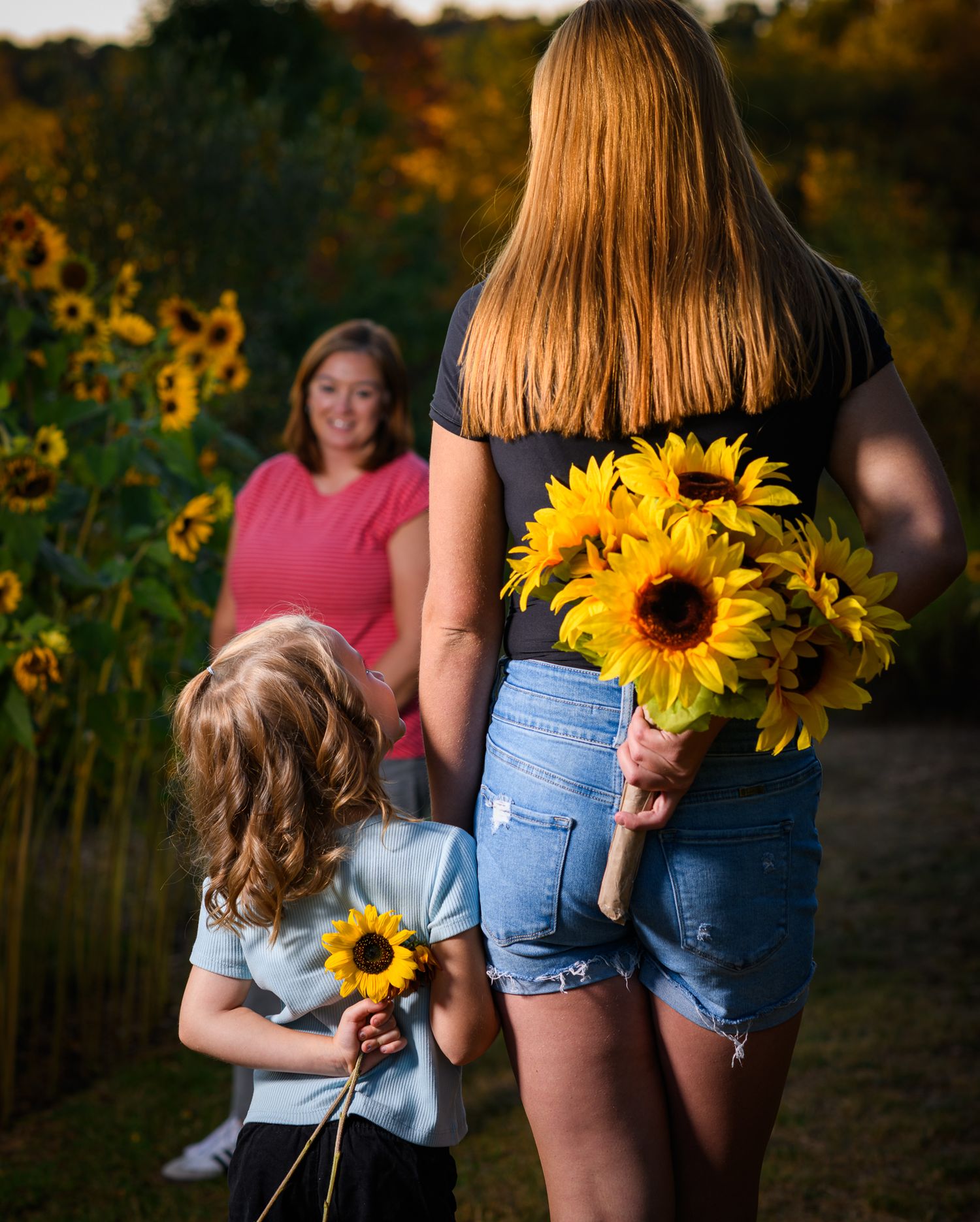Two girls holding sunflowers walk toward a smiling woman in a sunflower field during golden hour.