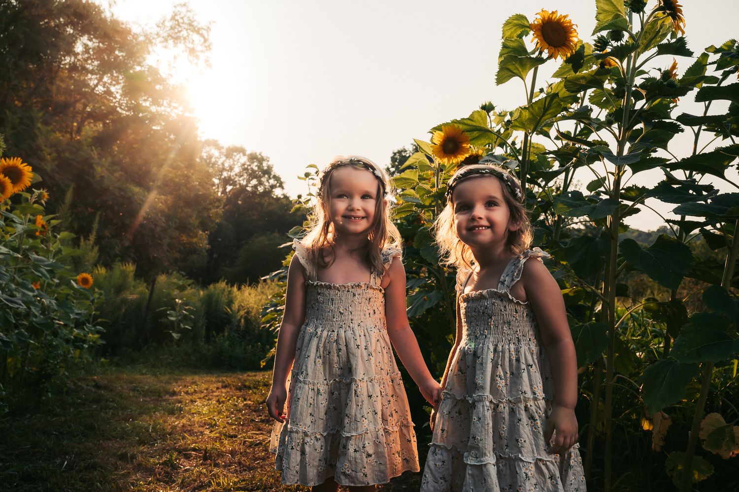 Two young girls in matching floral dresses hold hands among sunflowers in golden sunset light.