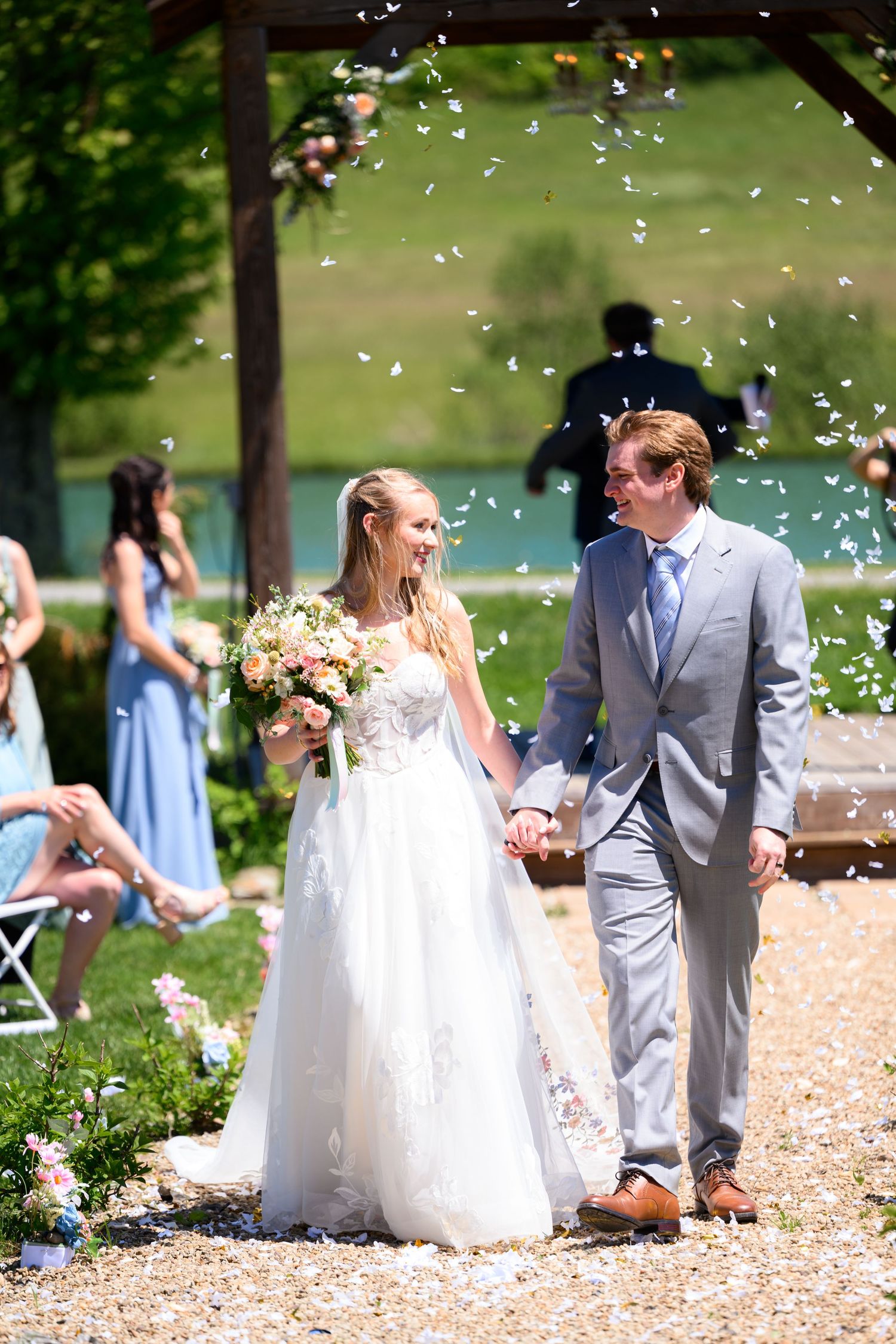 Bride and groom walk hand in hand as confetti falls during outdoor wedding ceremony near a lake.