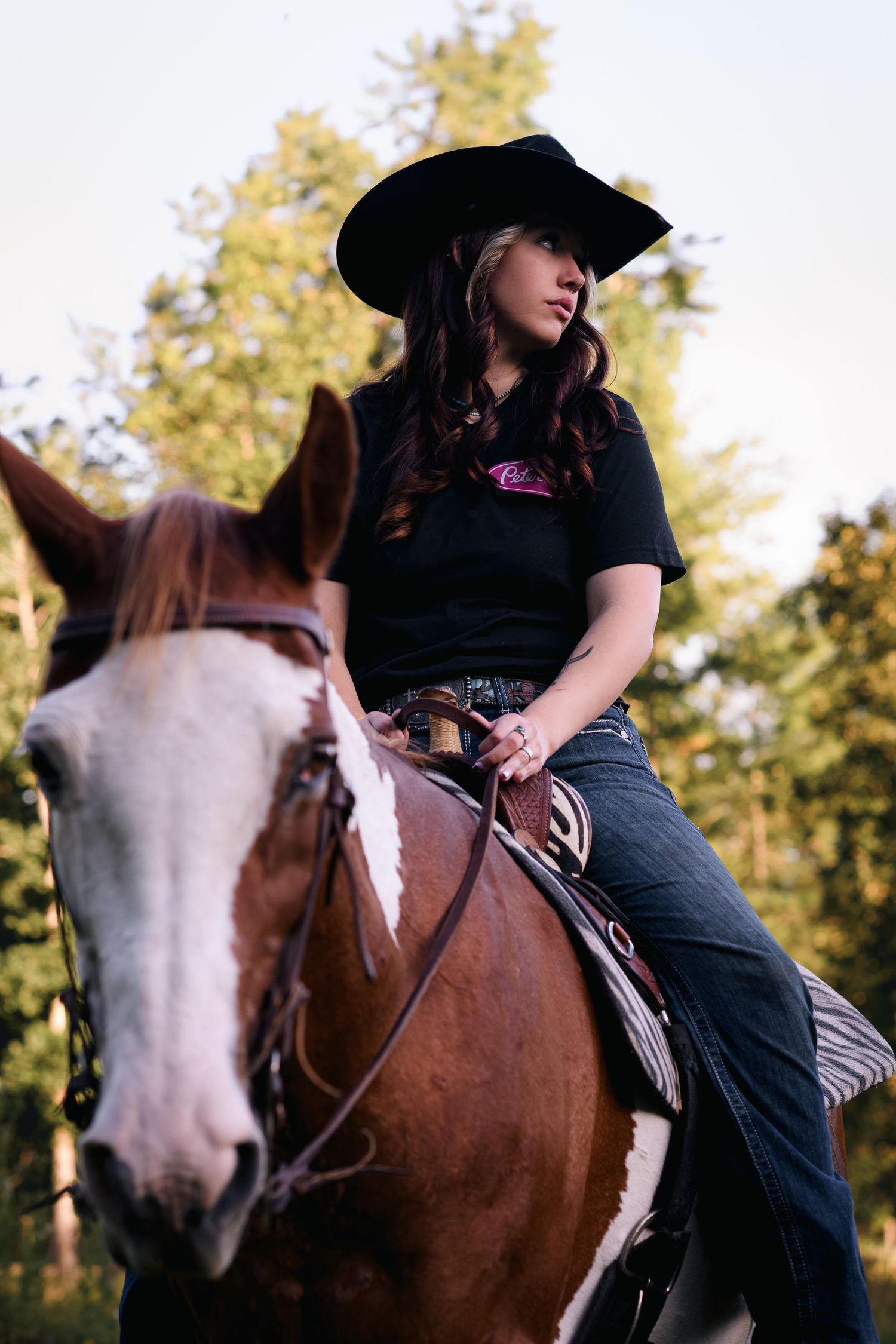 Young woman in black cowboy hat and jeans riding a brown and white paint horse outdoors.