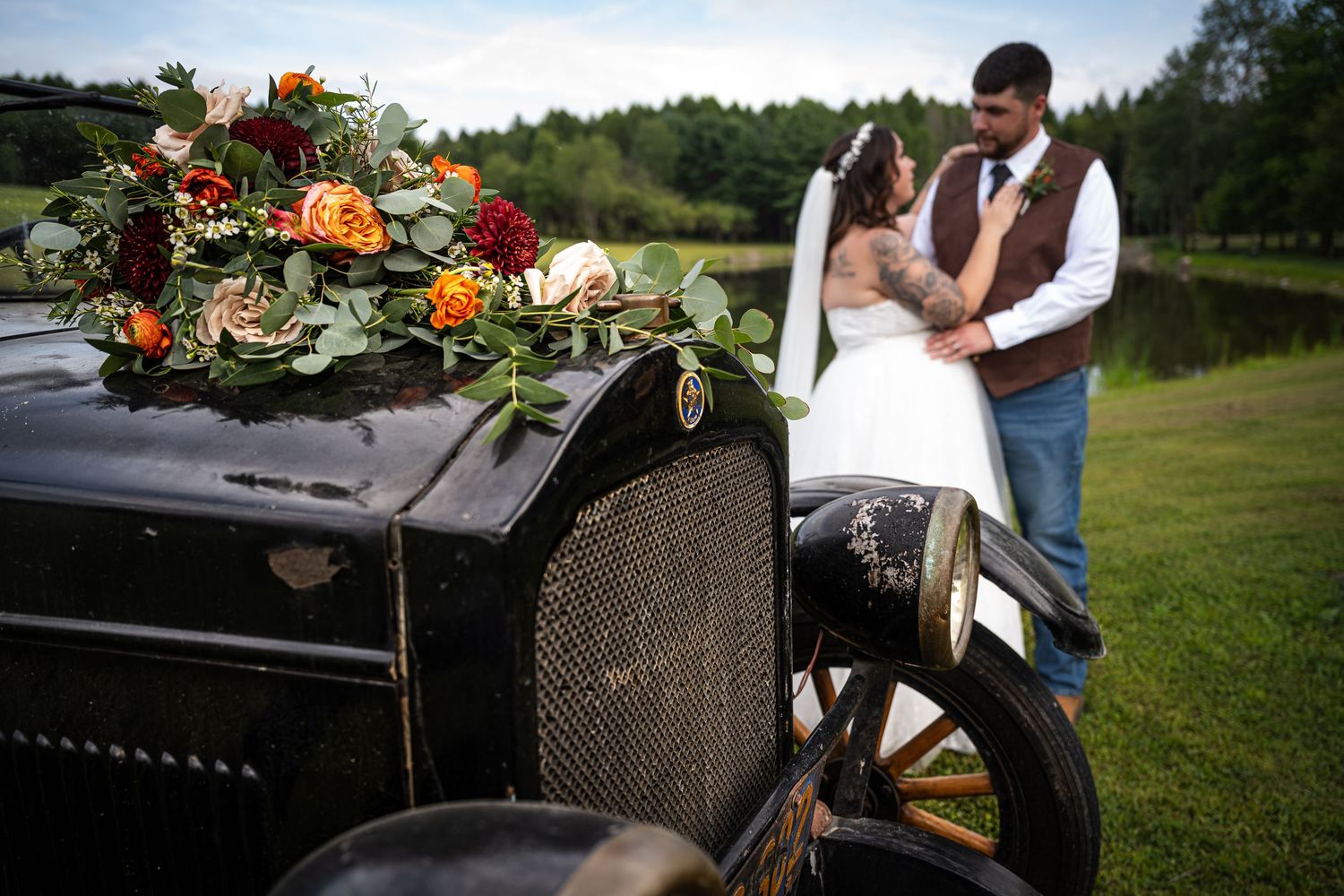 Bride and groom embrace beside vintage antique car adorned with lush floral wedding bouquet.