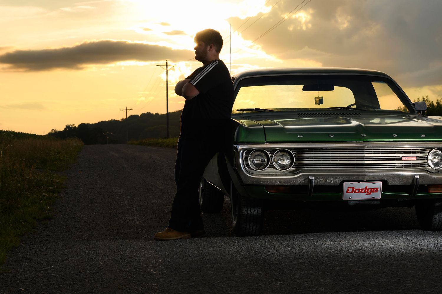 Man in suit leaning against a vintage green Dodge car on a rural road at sunset.