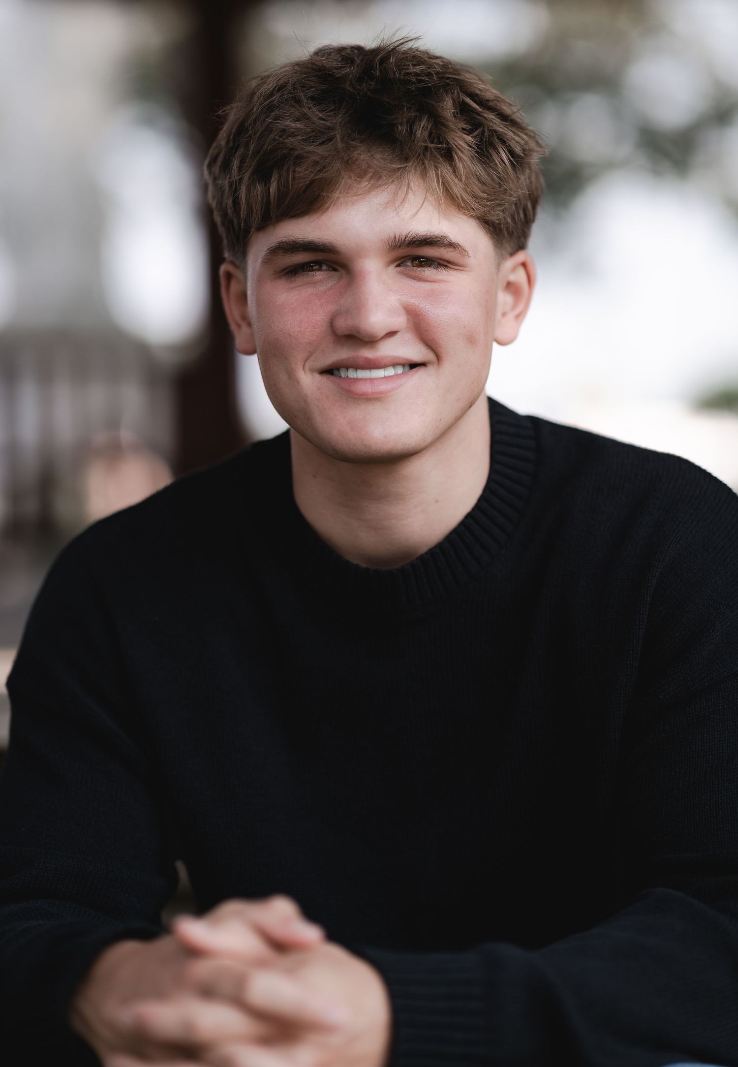 Young man in black sweater smiling with clasped hands in an outdoor portrait photo.