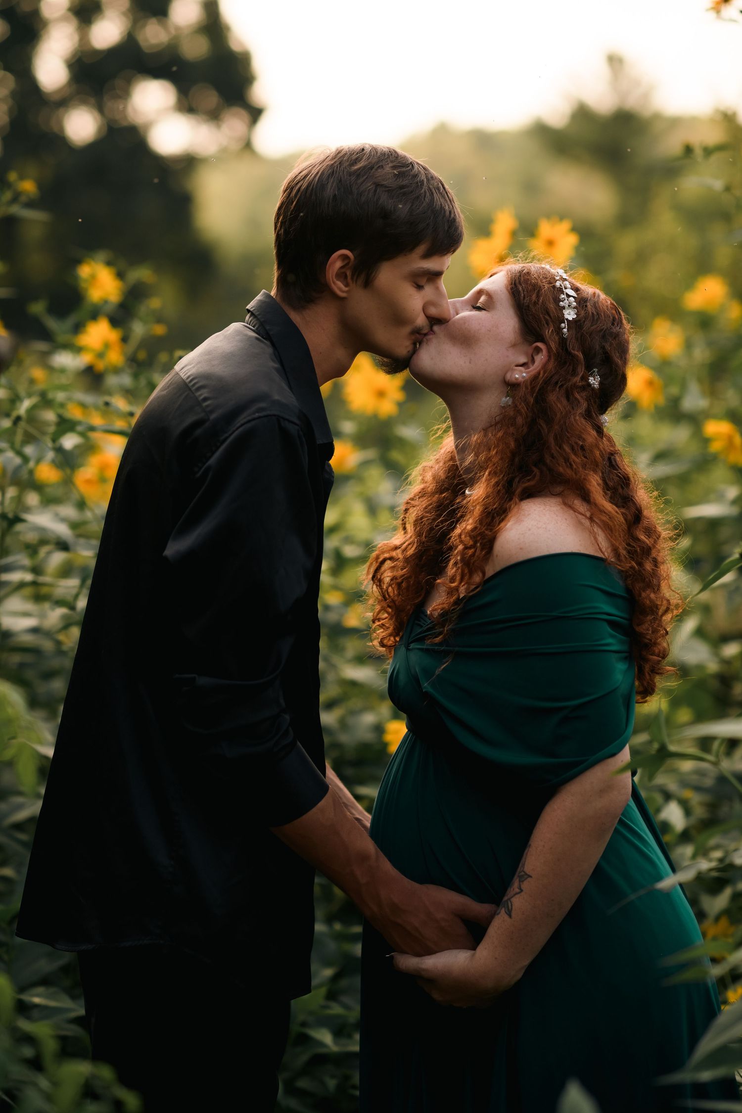 Couple kissing in sunflower field, woman in green off-shoulder dress with red hair, man in black shirt, golden hour.