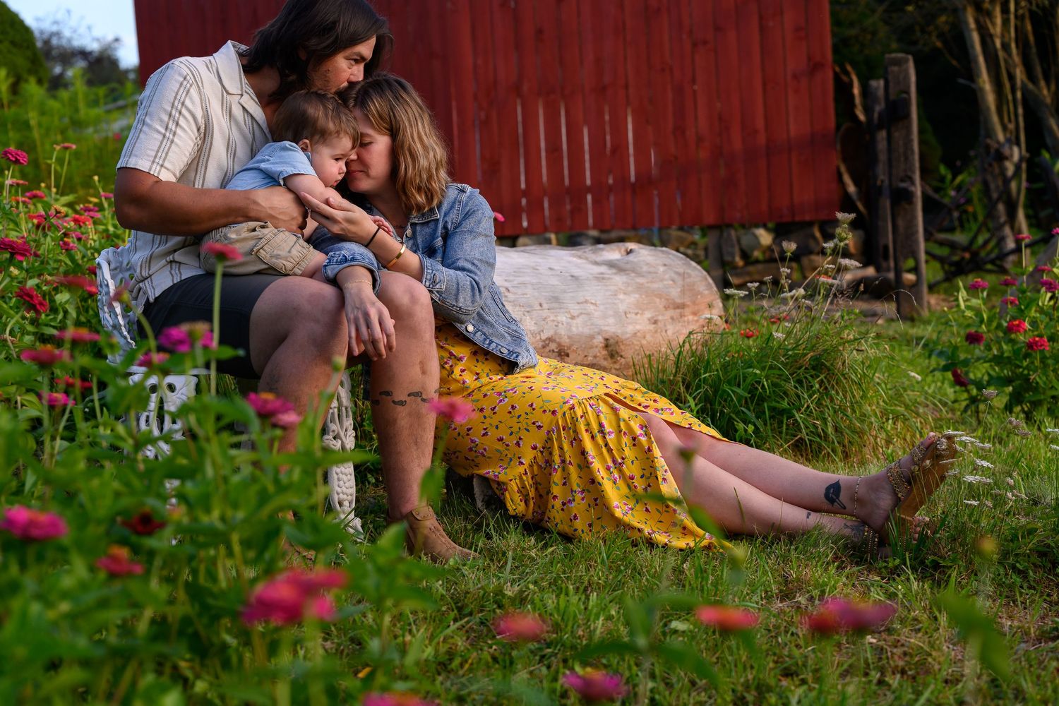 A family of three sits together in a lush garden near a red barn, sharing a tender embrace outdoors.