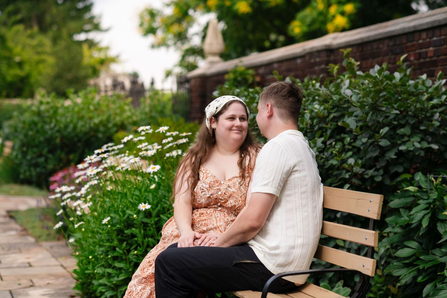 Couple sitting on a wooden bench in a garden surrounded by white flowers and lush greenery, smiling at each other.