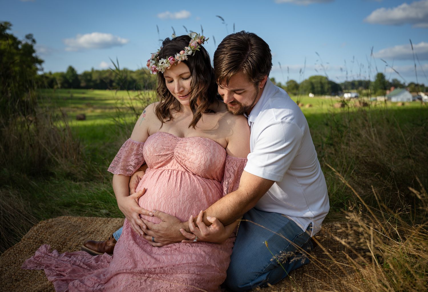 Couple sitting in field, man tenderly touches pregnant woman's belly as she wears pink gown.