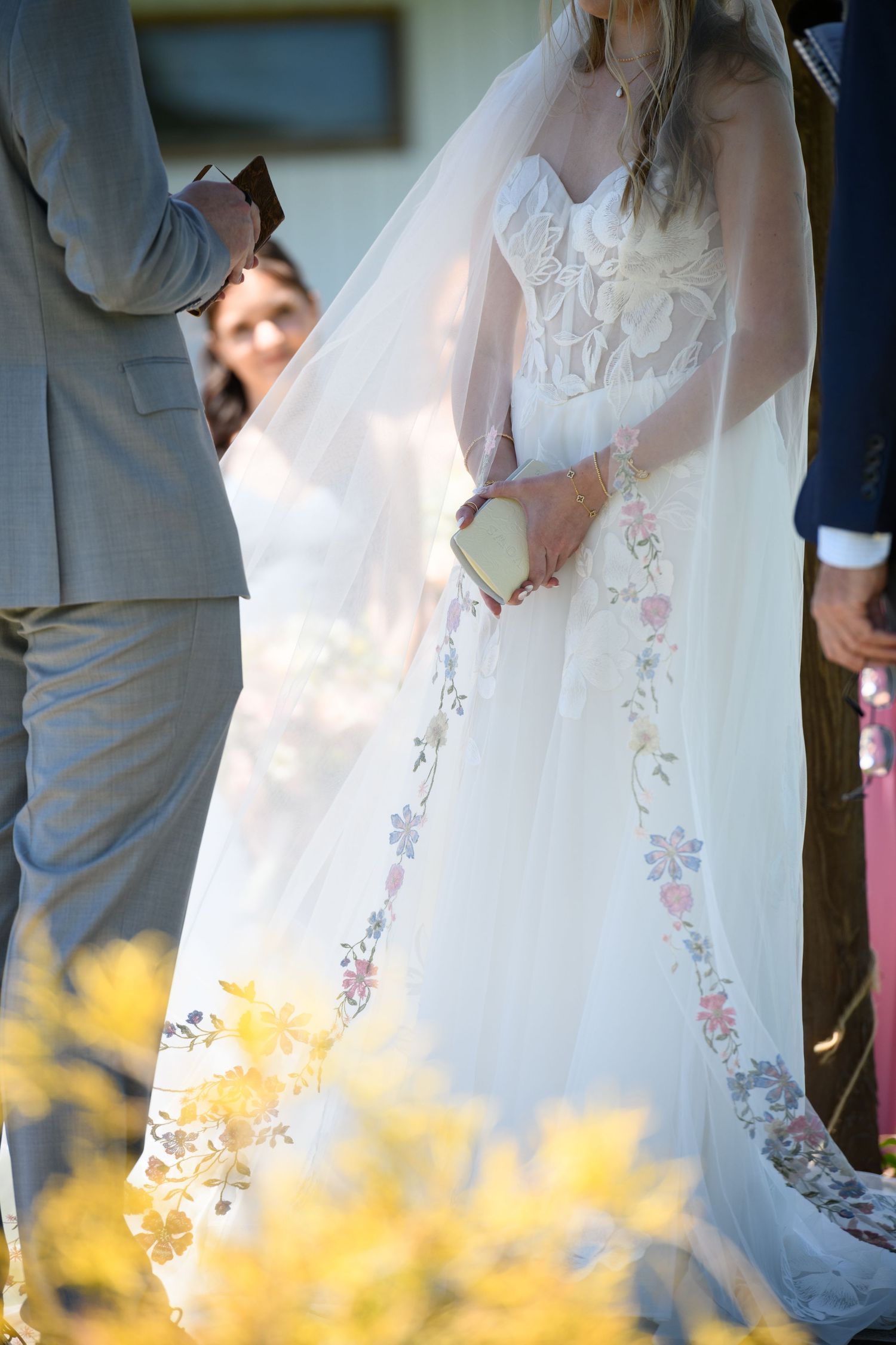 Bride in floral embroidered wedding gown with veil holds a small clutch during outdoor wedding ceremony.