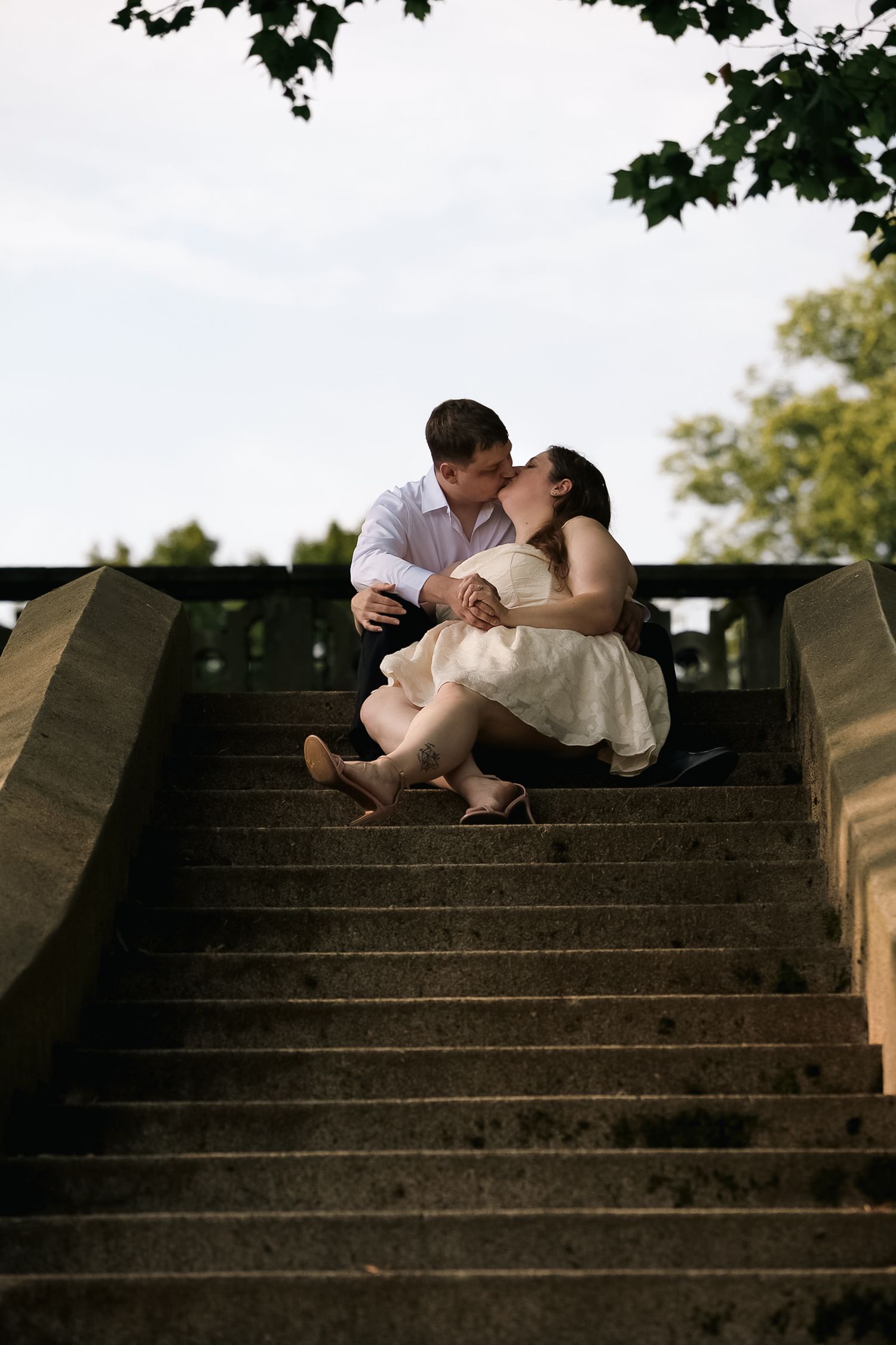 A couple shares a romantic kiss while sitting on outdoor stone steps, surrounded by lush green trees.