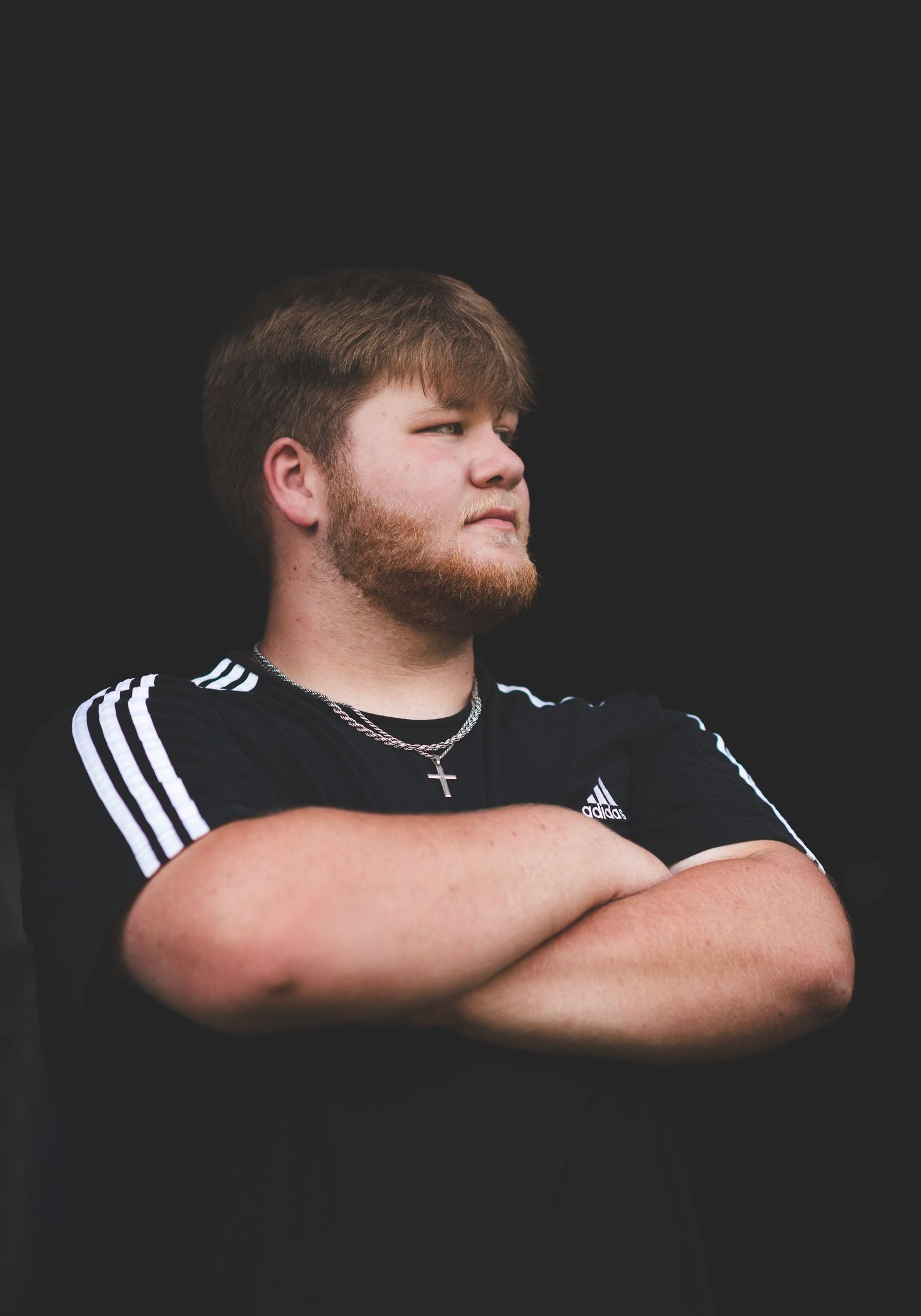 Young man in black Adidas shirt with arms crossed, wearing a cross necklace, posing confidently against dark background.