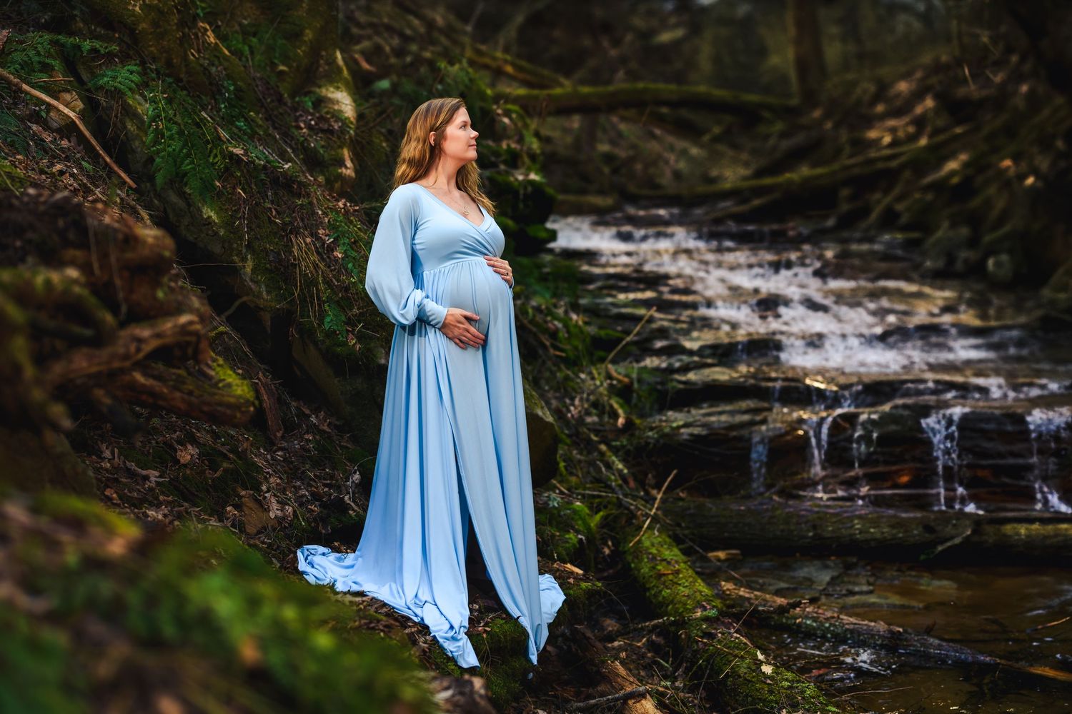 Pregnant woman in flowing light blue gown standing by a woodland waterfall, cradling her baby bump.