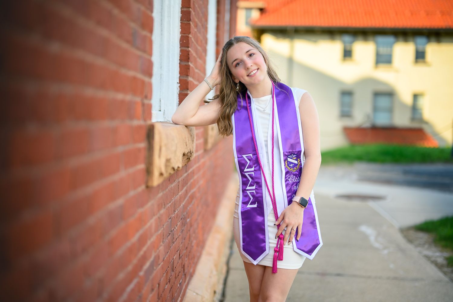 Smiling graduate in white dress with purple sash leans against brick wall outdoors.