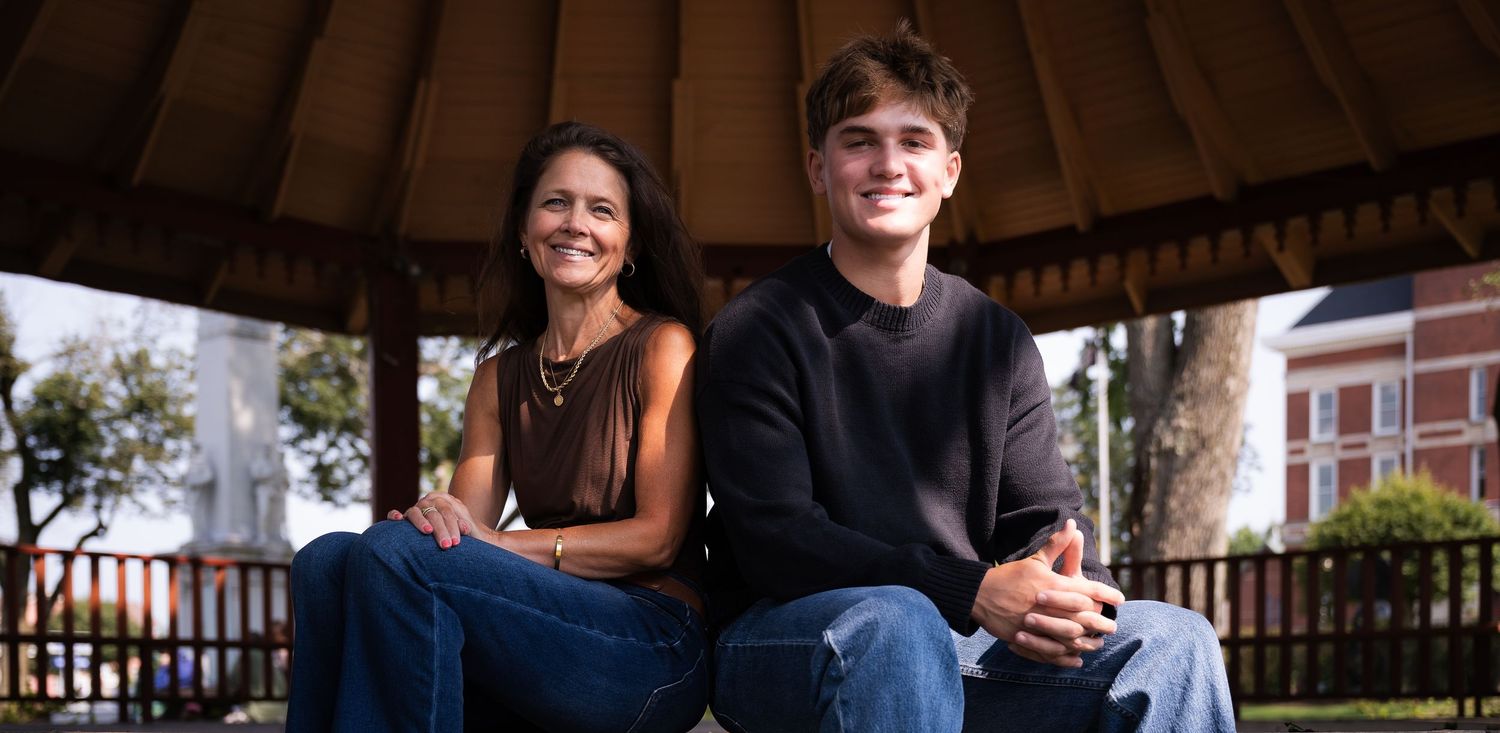 Two smiling people sitting together under a wooden gazebo on a sunny day outdoors.