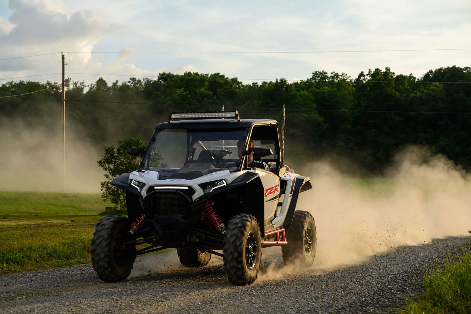 A white Polaris RZR off-road UTV kicks up dust on a gravel trail surrounded by green trees.