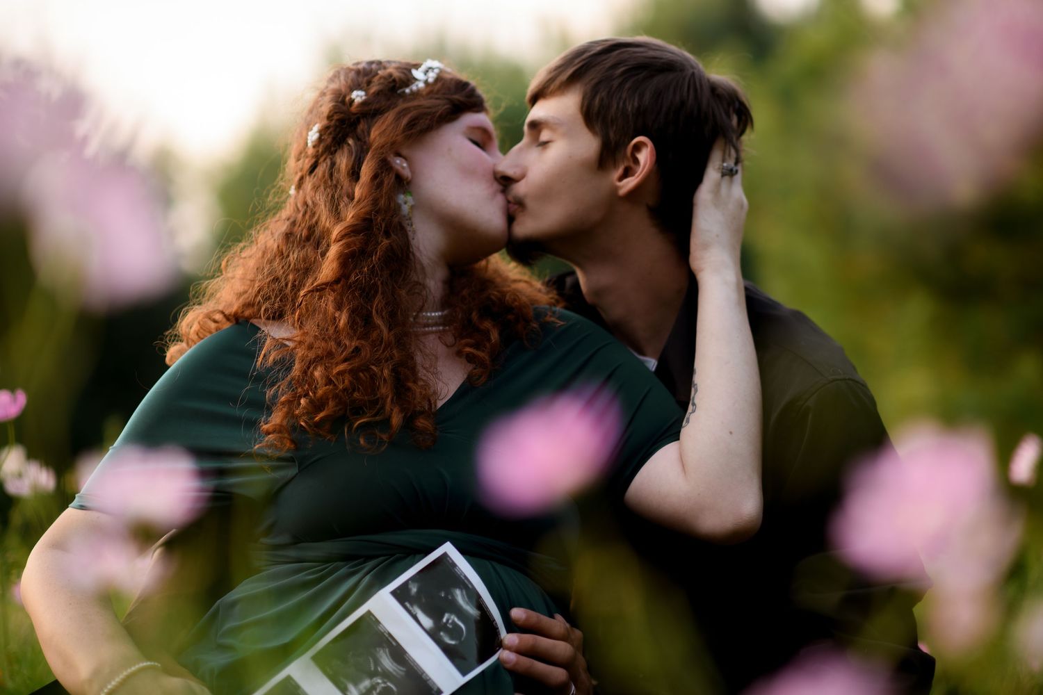 Couple kissing outdoors holding ultrasound photos, surrounded by pink flowers, announcing pregnancy.