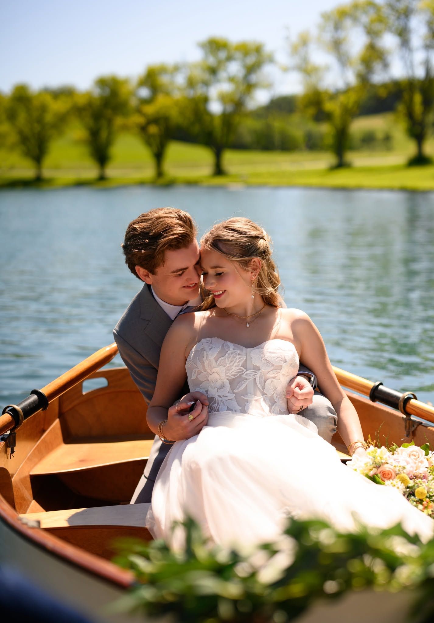 Romantic couple embracing in a wooden rowboat surrounded by flowers on a scenic lake.