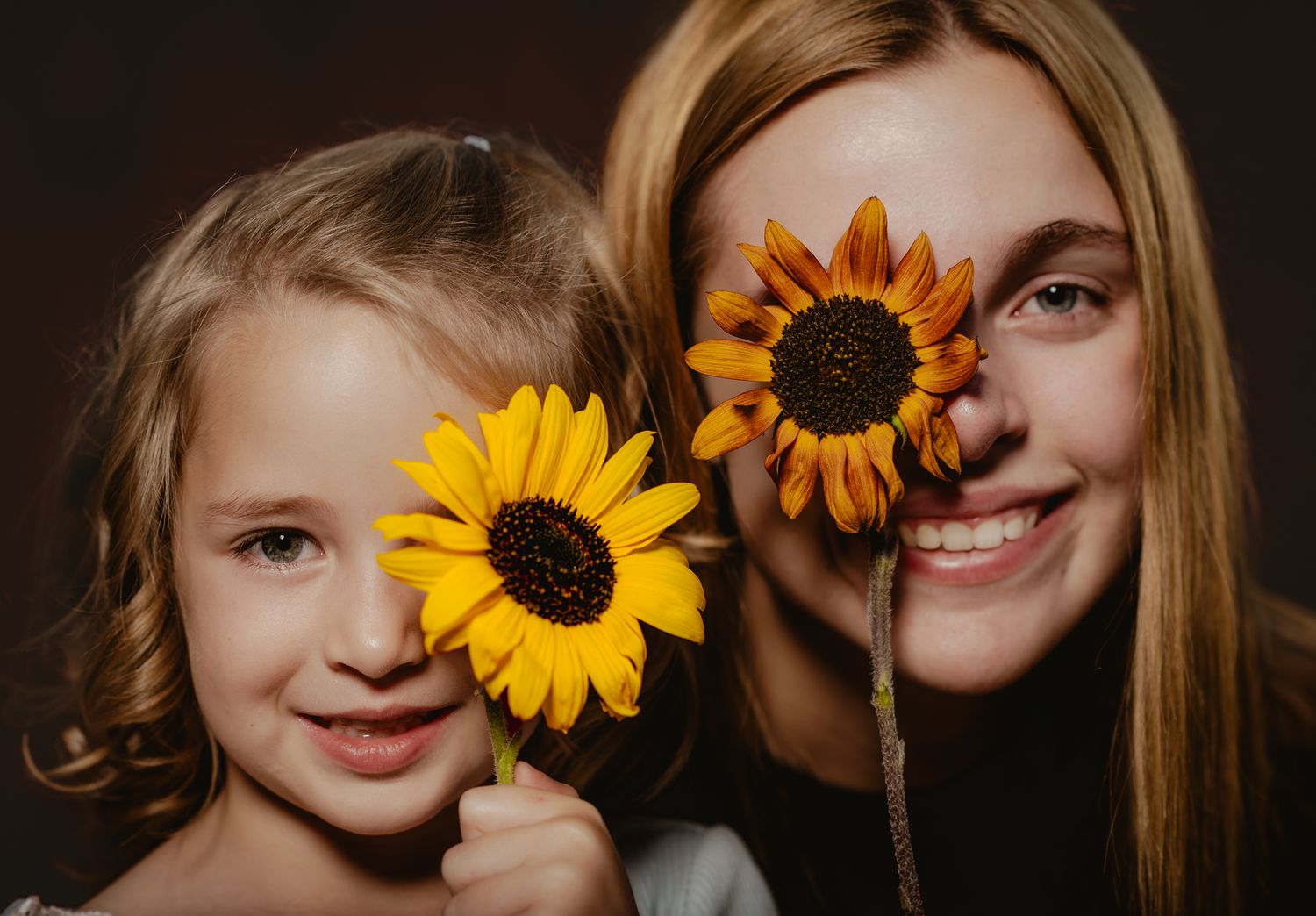 A smiling woman and young girl hold sunflowers over their eyes against a dark brown background.