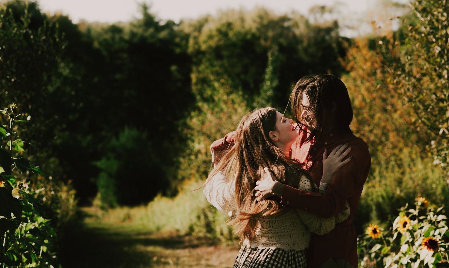 Two women share a tender embrace outdoors surrounded by lush green trees and sunflowers on a warm sunny day.