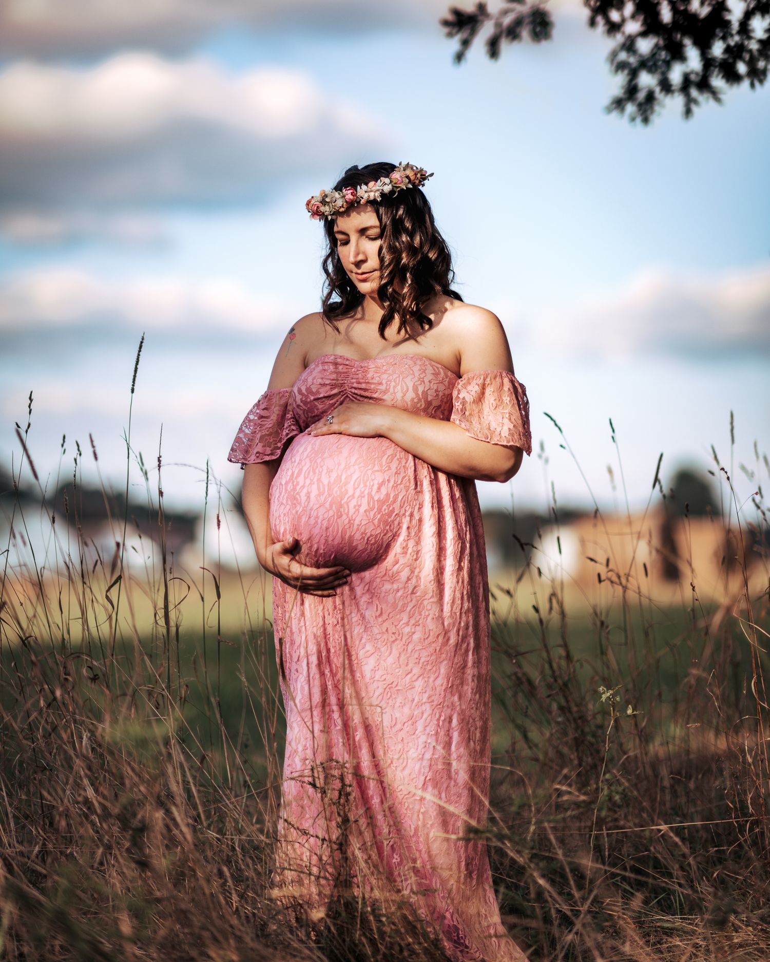 Pregnant woman in pink lace off-shoulder gown and floral crown stands in tall grass field.