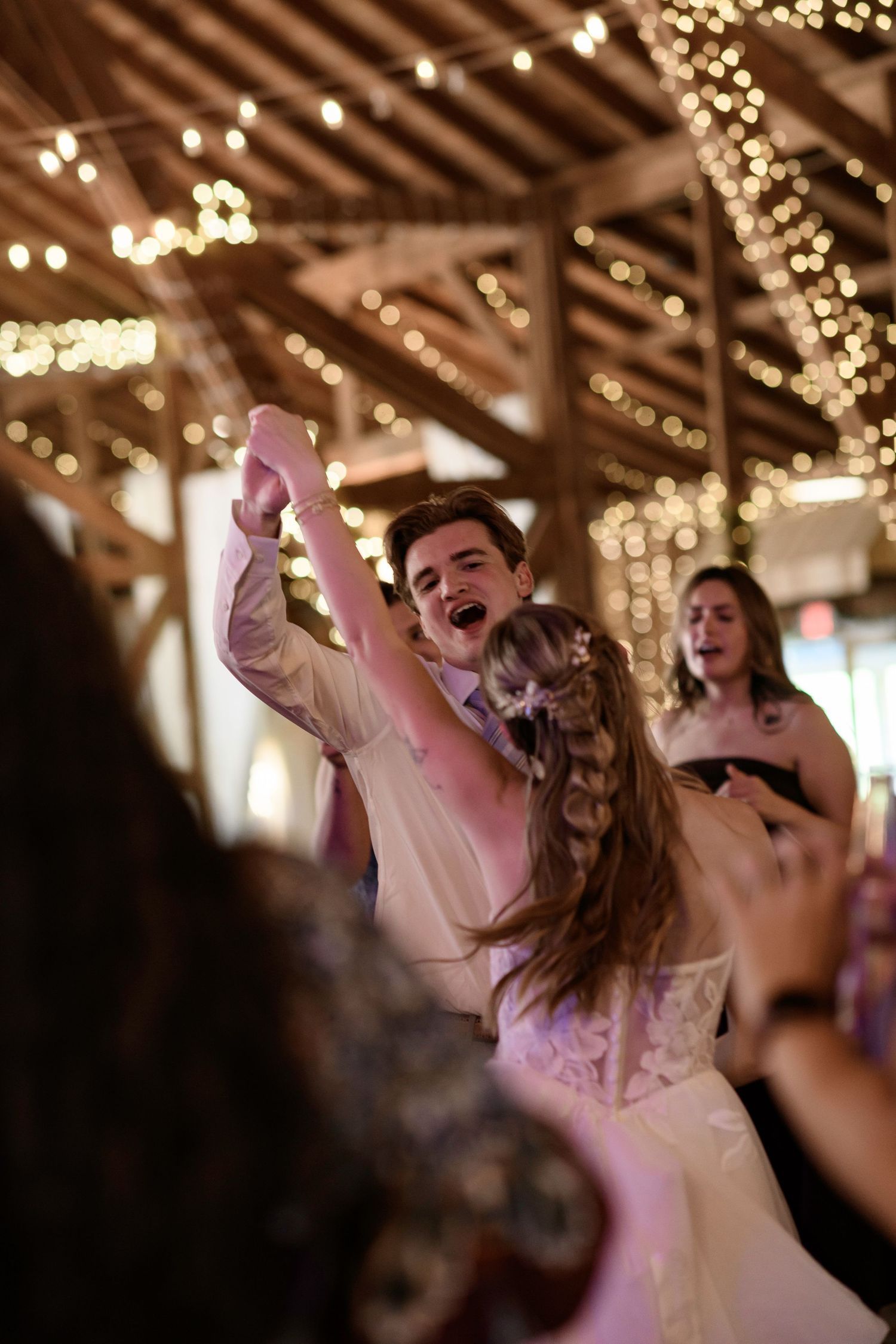 Bride and groom dancing joyfully at rustic barn wedding reception with warm string lights overhead.