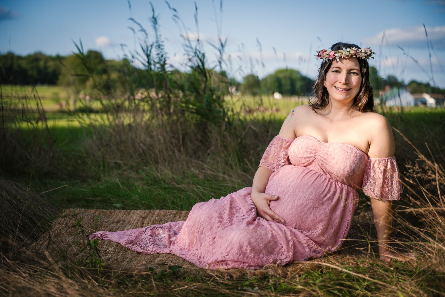 Pregnant woman in pink lace off-shoulder gown and floral crown sits in golden meadow for maternity photo.