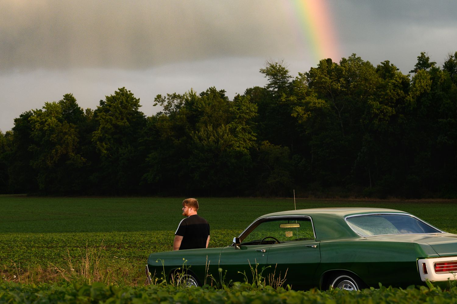 Man sitting on green classic car in field with rainbow and trees in background.