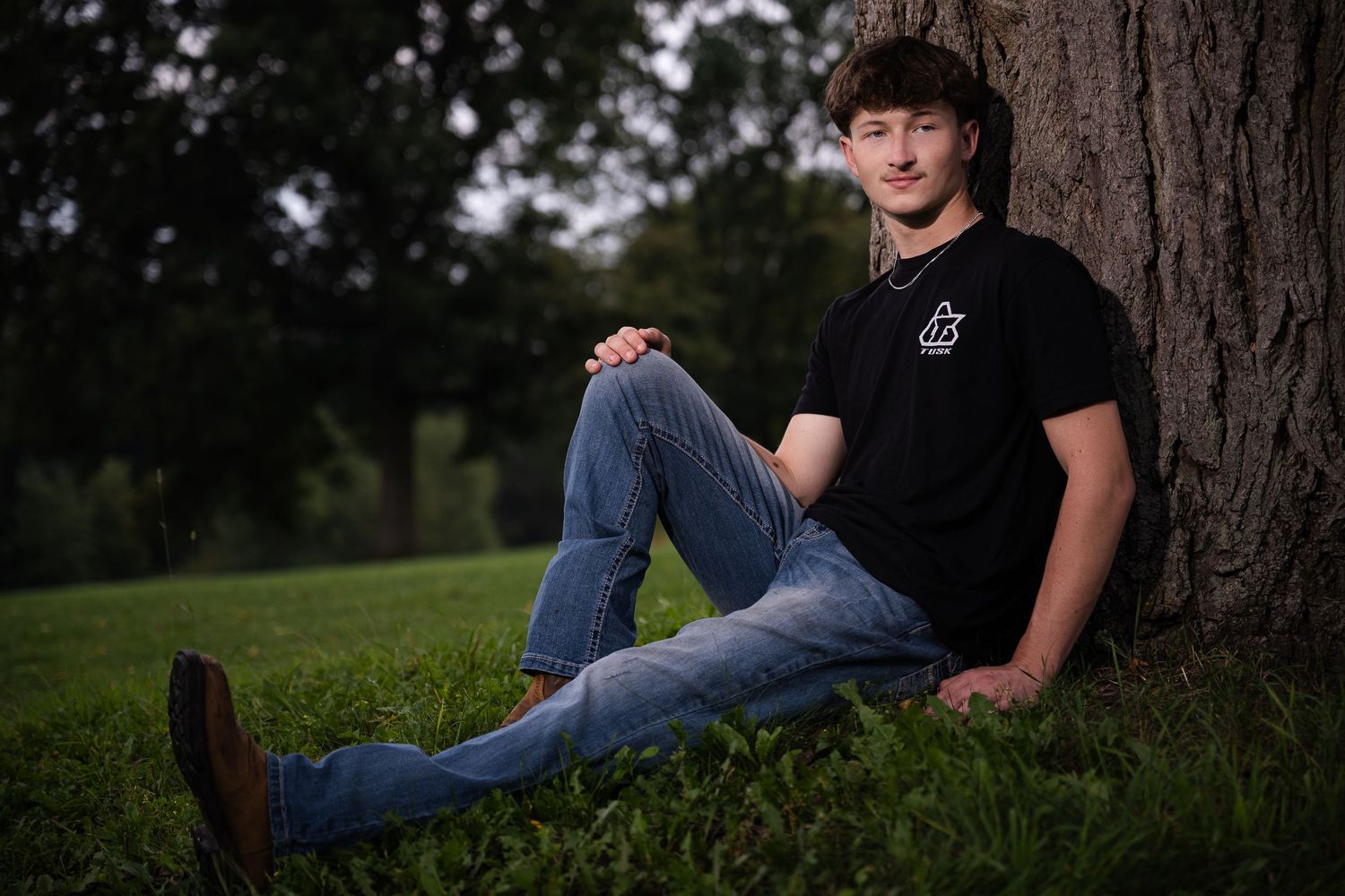 A young man in a black t-shirt and jeans sits relaxed against a large tree trunk on green grass outdoors.
