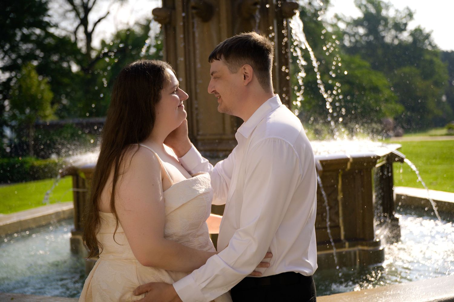 Couple sharing a romantic moment in front of a stone fountain in a lush green park on a sunny day.