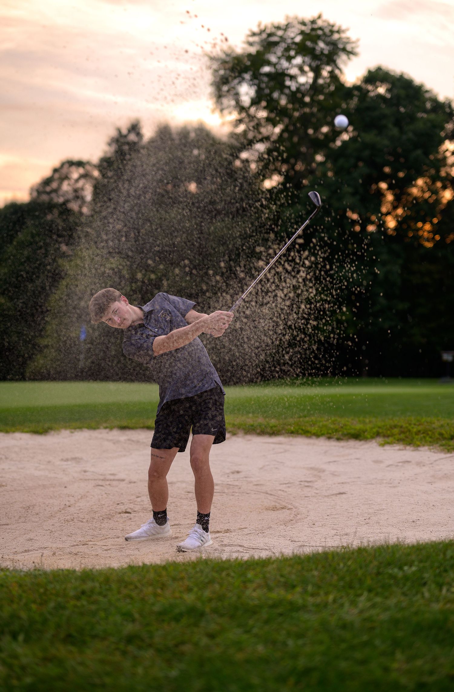 Young golfer hitting a sand bunker shot at sunset, sending sand flying on a lush golf course.