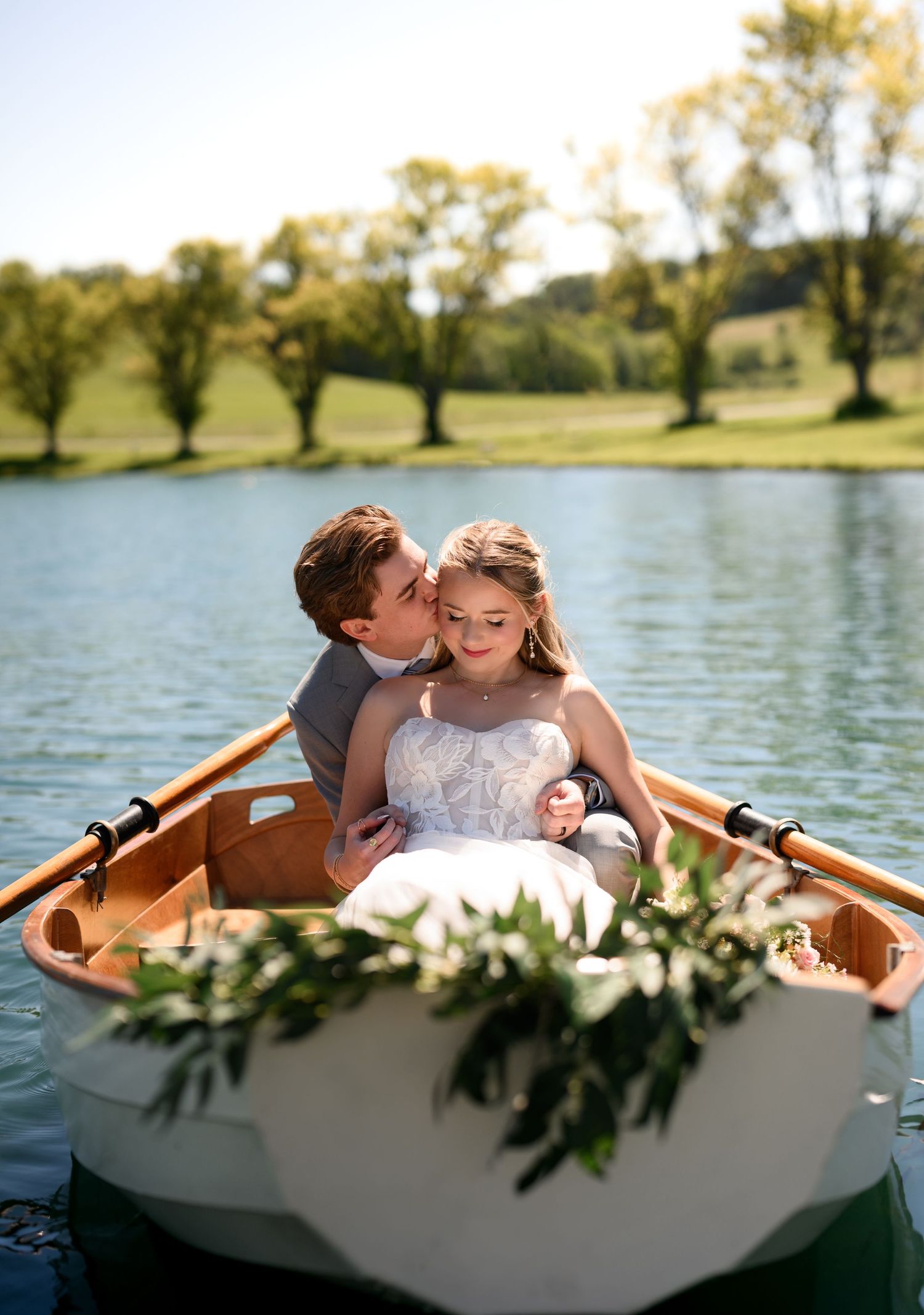 Bride and groom sharing a romantic kiss in a decorated rowboat on a serene lake surrounded by lush greenery.