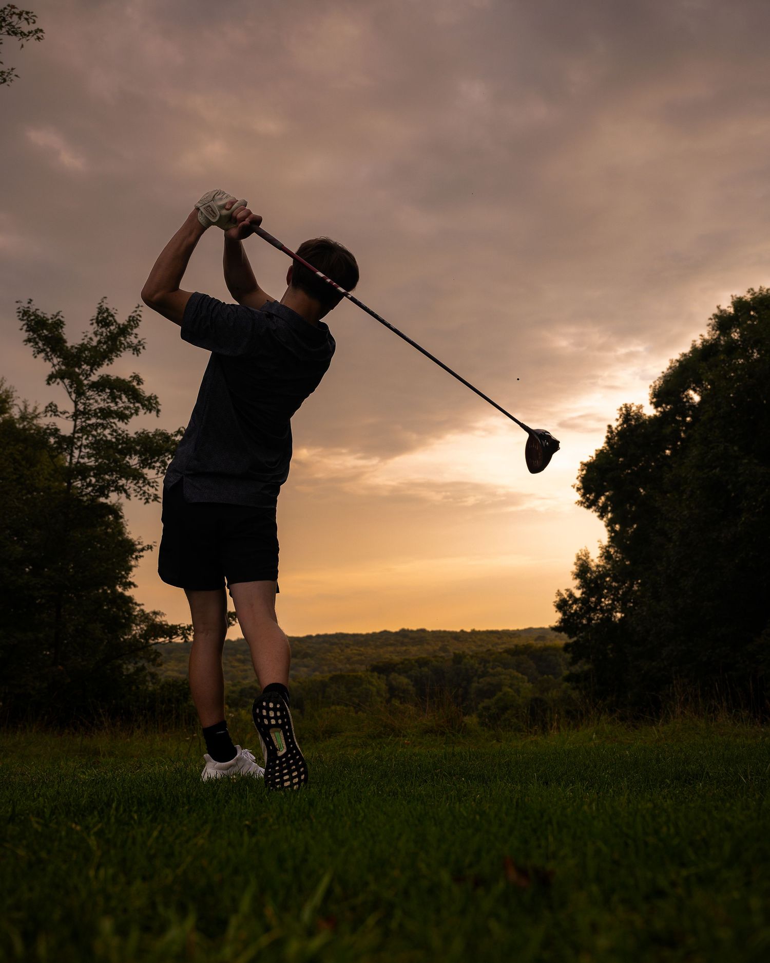 Golfer silhouetted against a dramatic sunset sky completing a full swing on a lush green golf course.
