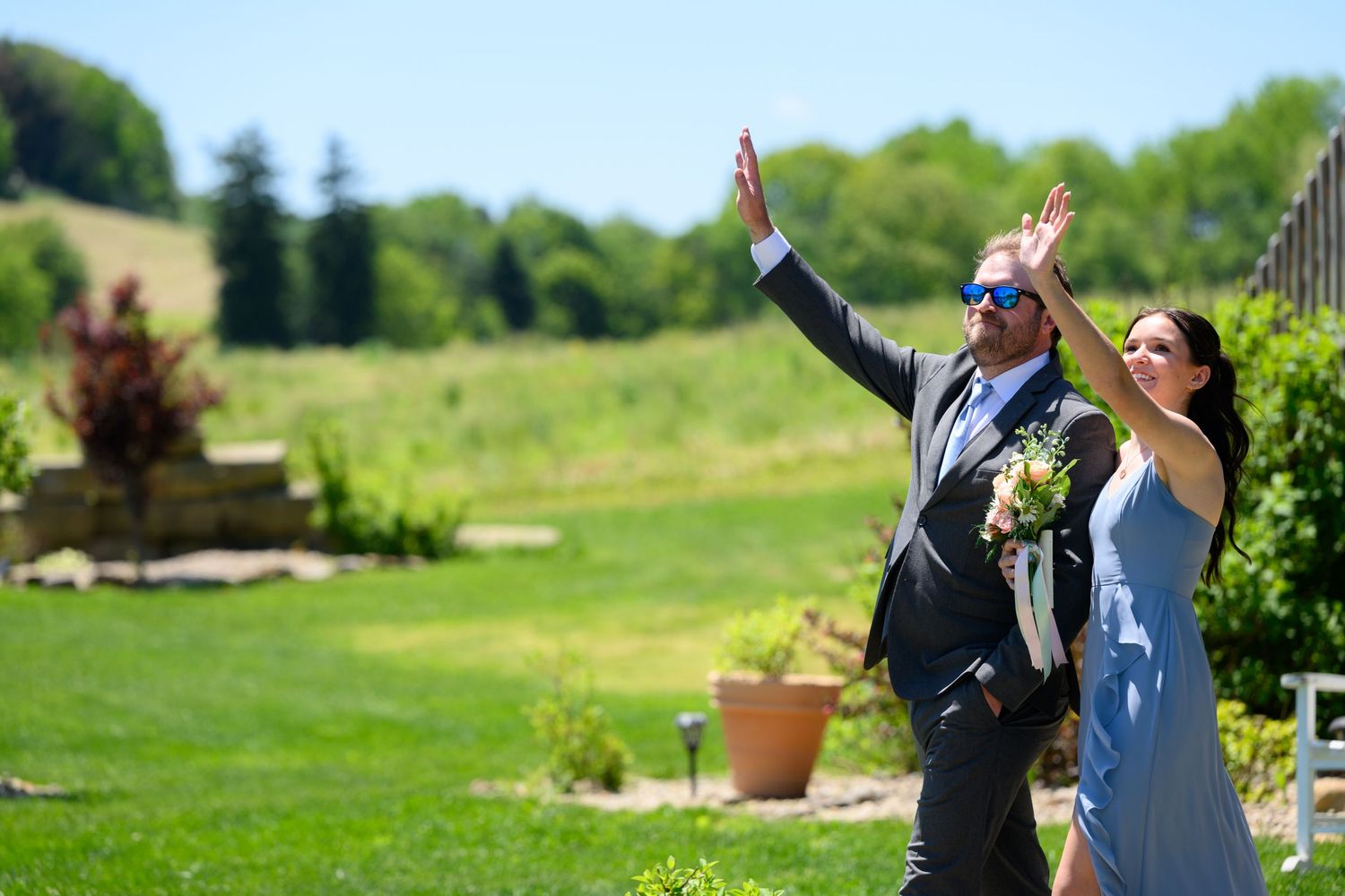 Wedding guests in blue attire wave and celebrate outdoors on a sunny day during a garden wedding reception.