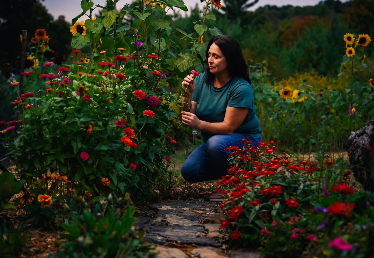 A woman crouches among vibrant red and pink flowers in a lush garden path.