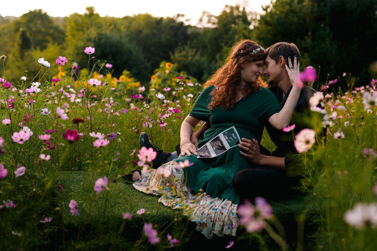 Couple sitting in a blooming flower field at sunset, holding ultrasound photos in a lush garden.