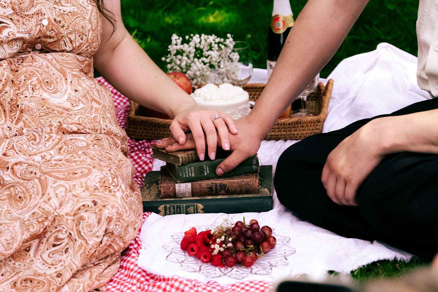 Couple holding hands at a romantic picnic with books, fresh berries, and a wicker basket on a floral blanket.