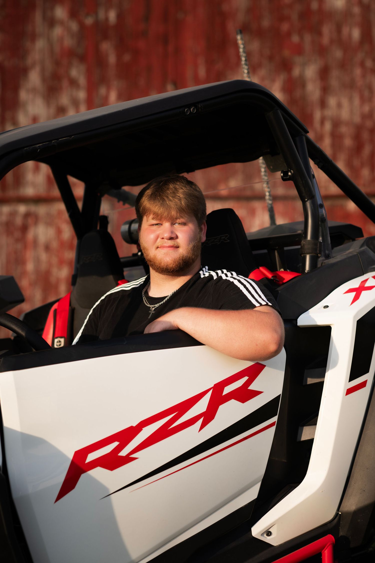 Young man sitting in a white Polaris RZR side-by-side UTV, wearing an Adidas shirt, posed in front of a red barn.