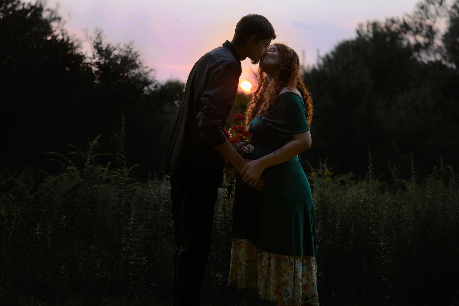 Couple sharing a romantic moment at sunset in a lush green field, silhouetted against a colorful twilight sky.