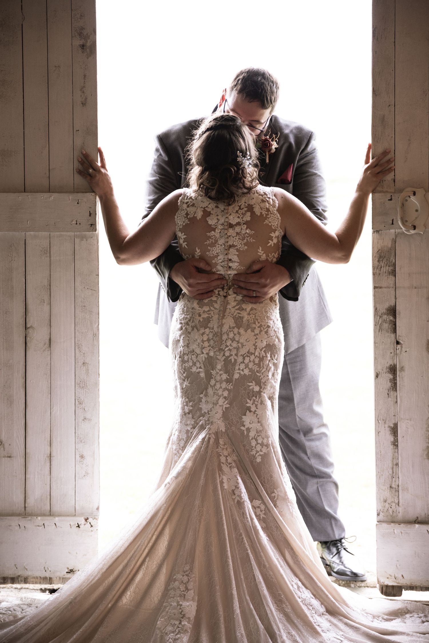 Bride in lace mermaid gown and groom share intimate kiss framed by rustic white barn doors.