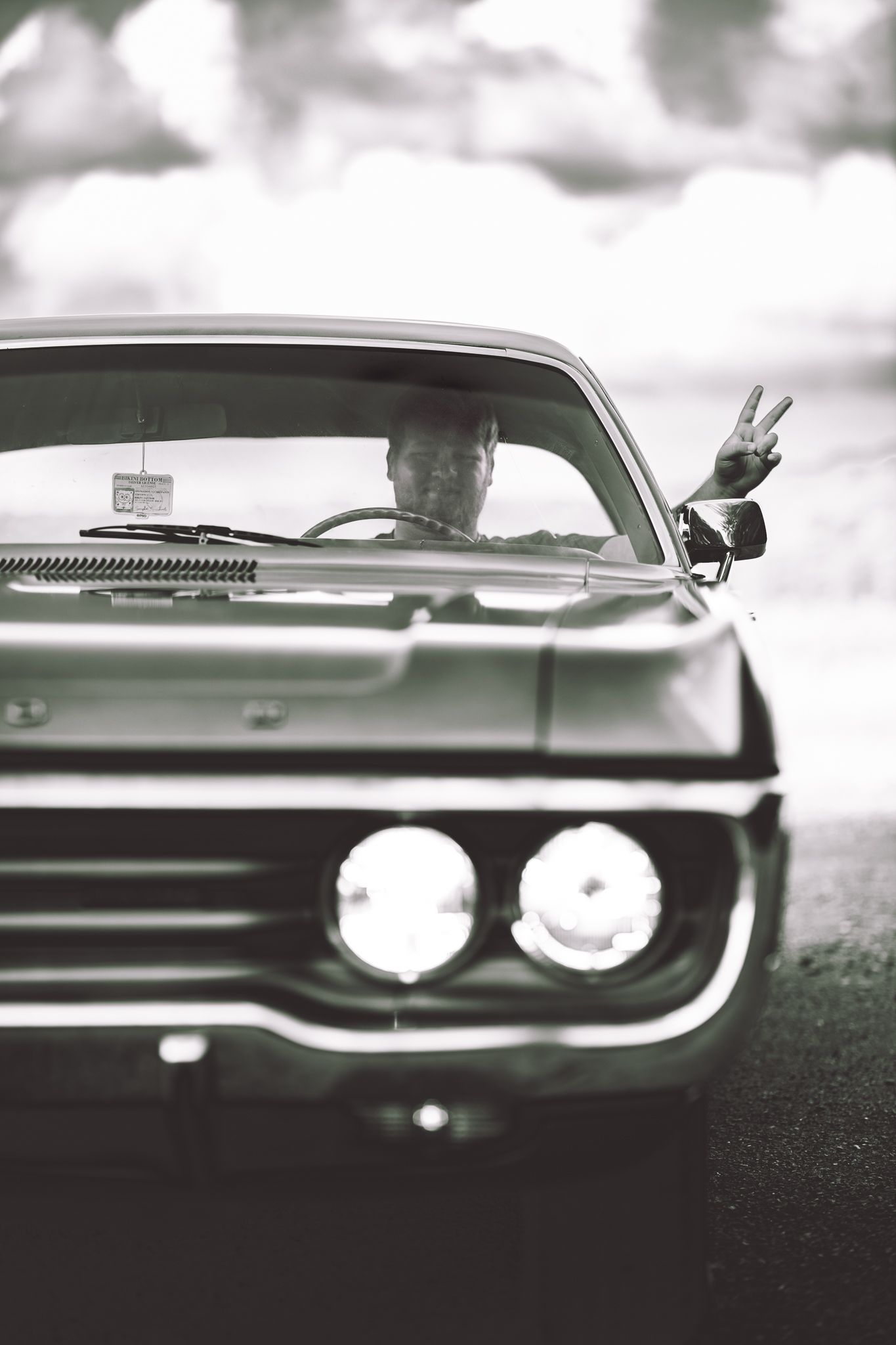 Black and white photo of a person flashing a peace sign from inside a classic vintage muscle car.