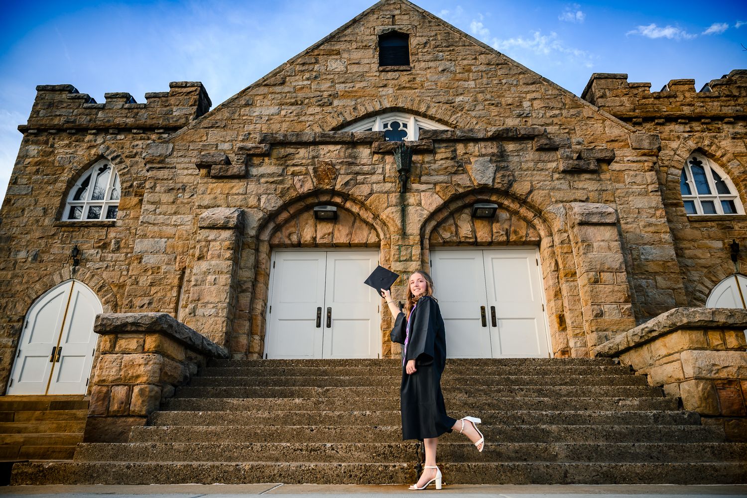 A graduate in cap and gown poses on steps before a grand historic stone church building.