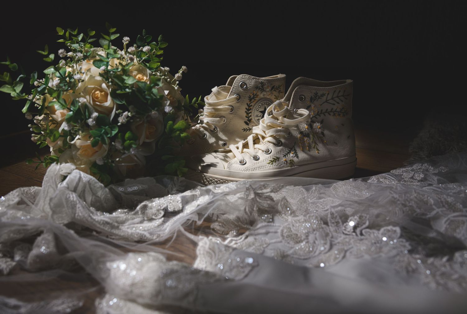 Decorated white high-top Converse wedding sneakers beside a floral bouquet and lace bridal veil on dark background.