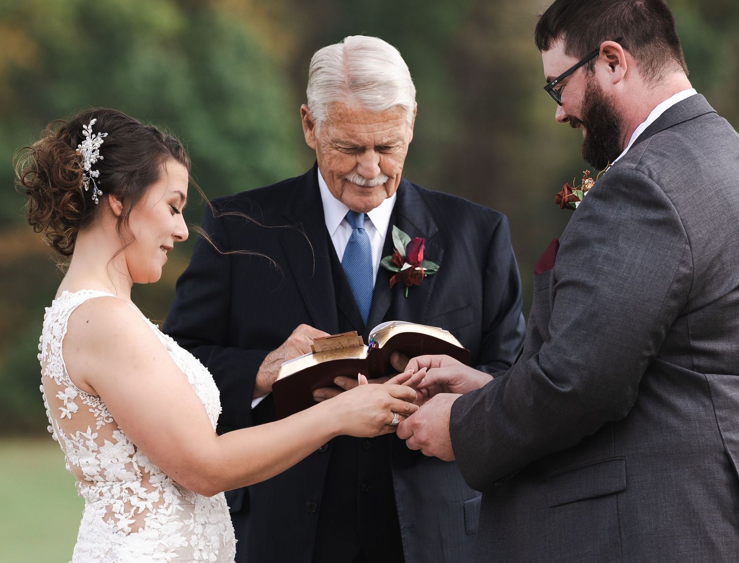 Bride places wedding ring on grooms finger as officiant reads from Bible during outdoor ceremony.